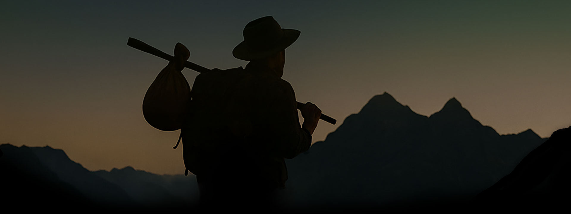 Silhouette of a vagabond with a backpack and bindle against a mountainous landscape during sunset